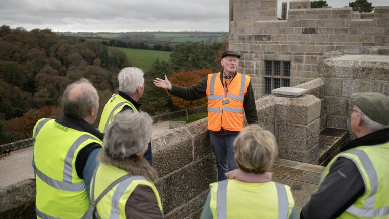 A guide in an orange hi-vis vest shows a group of visitors in yellow hi-vis around the roof at Castle Drogo, while explaining about the landscape and construction work.